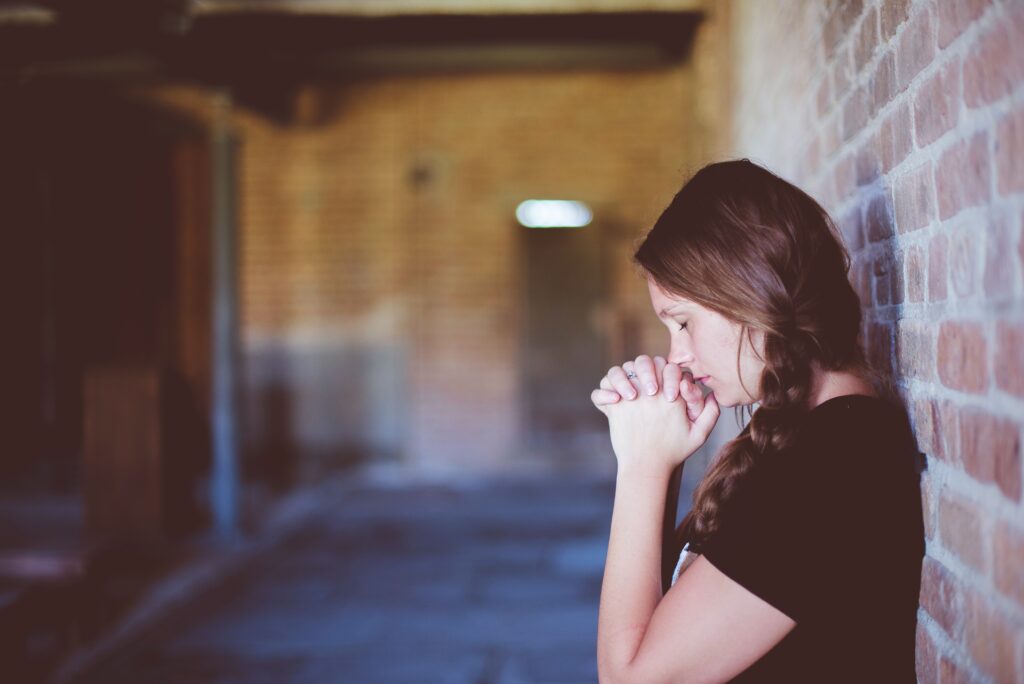 A young woman stands isolated in a brick building, her back leaning against a brick wall, with hands clasped at her lips with her head bowed.