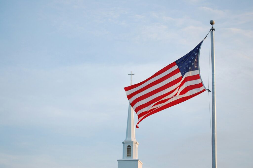 A church steeple topped with a cross behind a flying US flag.
