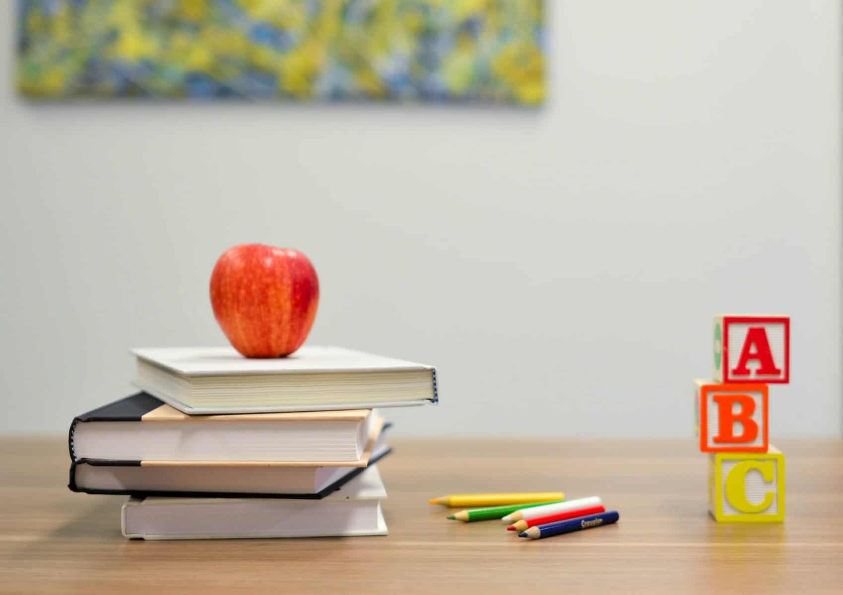A red apple sits on a stack of books on a table top with colored pencils and alphabet blocks stacked nearby.