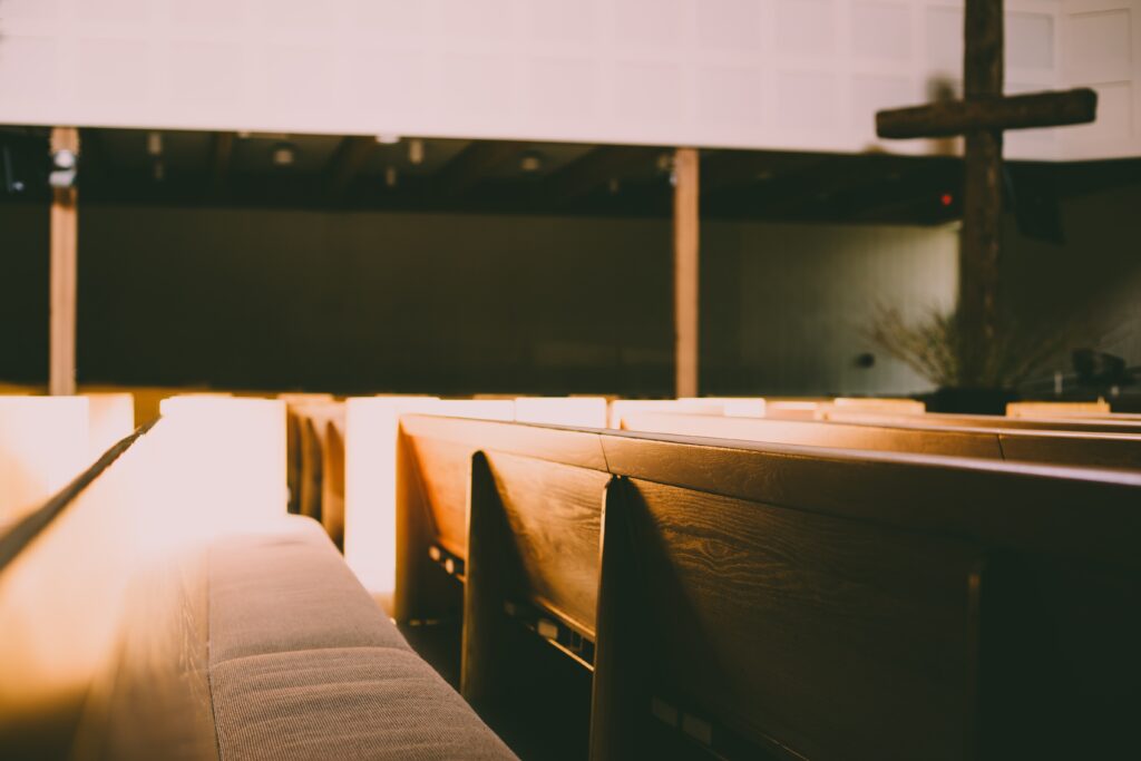 Light brown church pews are seen from the side in the foreground while a dark brown cross is seen at the top right of this horizontal image.