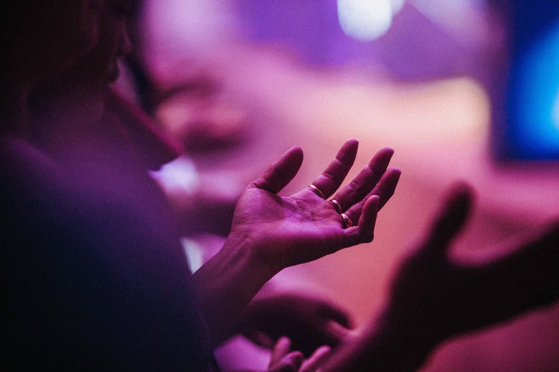 A woman's hand with gold rings on three fingers is lifted slightly in front of her, palm up in an atmosphere with pink and orange lights such as at a concert.