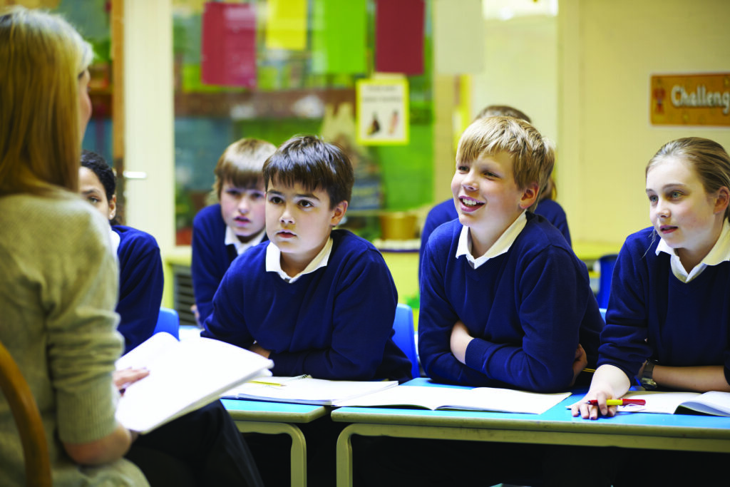 Three children are clearly seen facing the camera wearing white shirts and blue sweaters and leaning on a school table. The teacher is mostly out of frame.